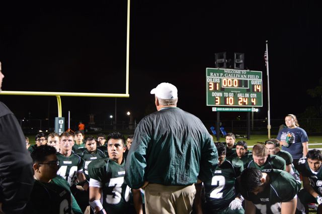 A coach addresses seated players on the field beneath the scoreboard.