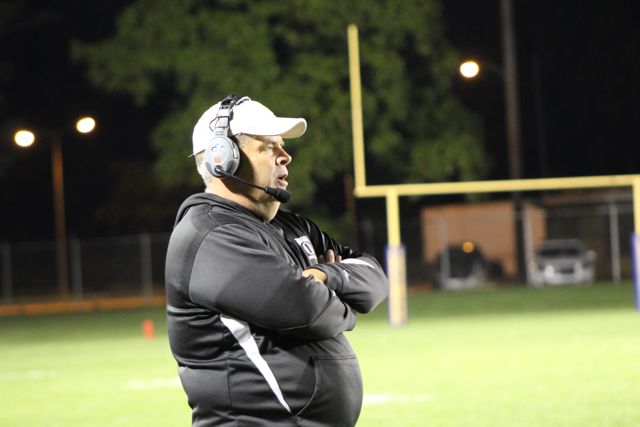 A coach wearing a headset watches the field under the lights.