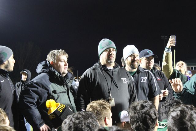A coach speaks to players and supporters gathered on the field.